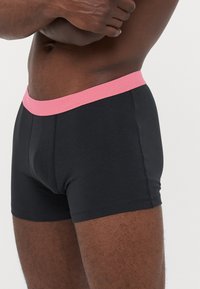 Nahaufnahme einer Person, die schwarze Boxer-Shorts mit einem rosa Bund trägt, die Arme über einem nackten Oberkörper verschränkt, vor einem einfarbigen Hintergrund.