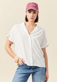 Camisa blanca de manga corta con botones y un patrón texturizado, combinada con jeans de color azul claro y una gorra rosa con el texto 'California'.