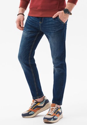 Mężczyzna w niebieskich obcisłych dżinsach, wielokolorowych trampkach, czerwonej bluzie, czarnym zegarku i czarnej bransoletce, stojący z jedną ręką w kieszeni.