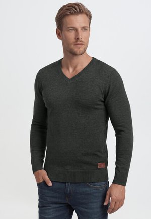 Homme aux cheveux châtain clair portant un pull en V gris foncé et un jean bleu, debout avec une main dans la poche sur un fond uni.