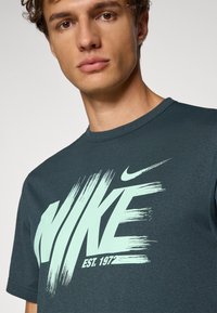Mørk teal kortærmet t-shirt med stort hvidt og mintgrønt "NIKE" logo i penselstrøgsdesign, rund halsudskæring.