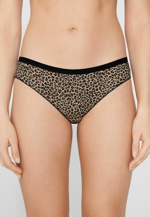 Vrouw die een bikini-ondergoed met luipaardprint en een zwarte tailleband draagt, staand tegen een effen lichte achtergrond.