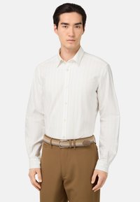 Camicia bianca a maniche lunghe con righe verticali, colletto button-down e tessuto morbido, abbinata a pantaloni marroni e una cintura intrecciata.