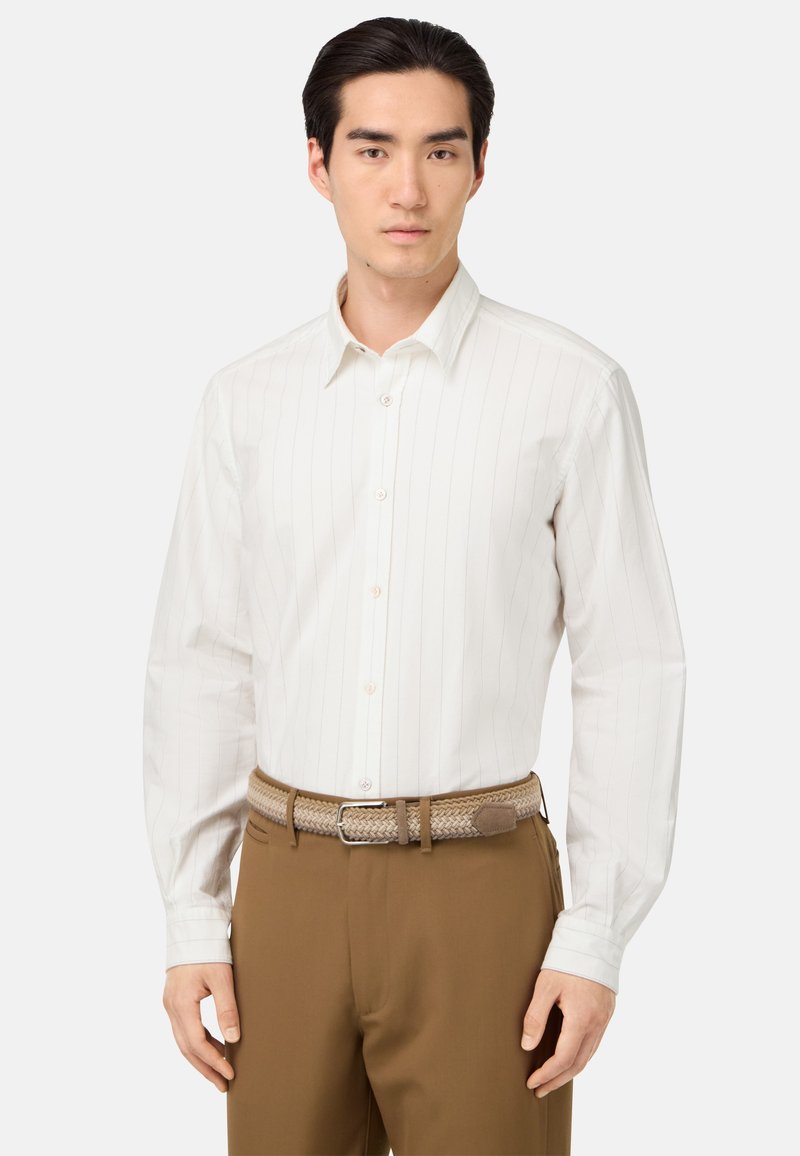 Camicia bianca a maniche lunghe con righe verticali, colletto button-down e tessuto morbido, abbinata a pantaloni marroni e una cintura intrecciata.
