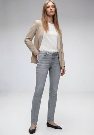 Donna in piedi che indossa un blazer beige, una t-shirt bianca, jeans grigio chiaro e ballerine nere su uno sfondo grigio chiaro uniforme.