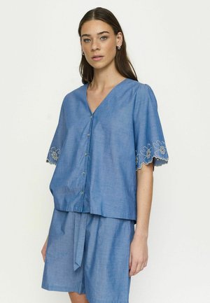 Fiatal nő kék gombos blúzban, virágmintás hímzett rövid ujjakkal és hozzá illő, bővebb rövidnadrágban, egyszínű háttér előtt.