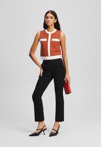 Brun teksturert crop topp med hvite detaljer, paret med svarte tettsittende bukser og svarte stilettosko. Modell holder en rød clutch.