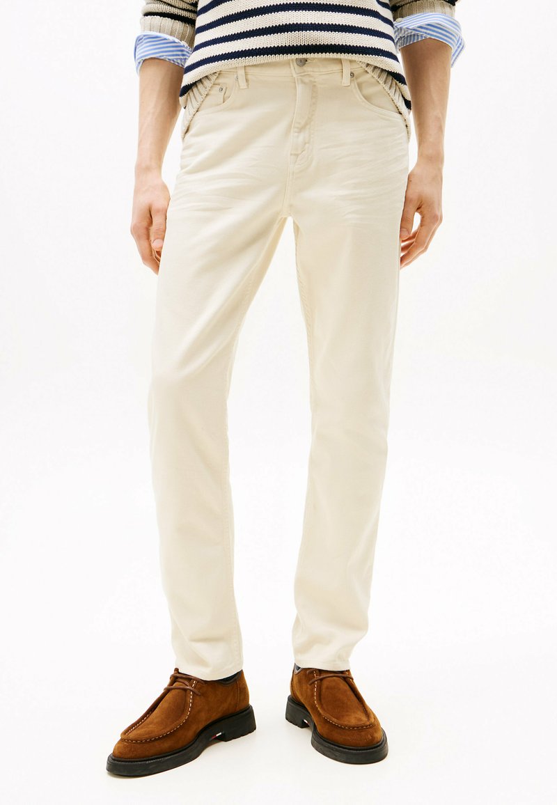 Pantalones de algodón beige con corte ajustado, que cuentan con bolsillos laterales y una cinturilla con pasadores para cinturón, combinados con zapatos de ante marrones.