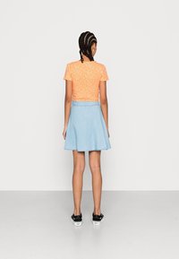 Orangefarbenes, kurzärmeliges Oberteil mit Blumenmuster, kombiniert mit einem hellblauen Denim-A-Linien-Rock und schwarzen Sneakers; geflochtene Frisur sichtbar.