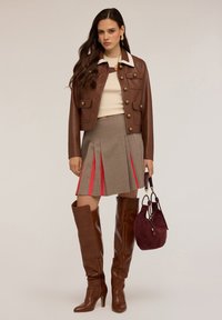 Donna con giacca di pelle marrone, top beige, gonna plissettata a quadri con inserti rossi, stivali marroni fino al ginocchio, che tiene una borsa borgogna.