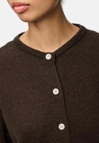 Cárdigan marrón de punto con cuello redondo, que presenta tres botones blancos y una textura acanalada en el collar.
