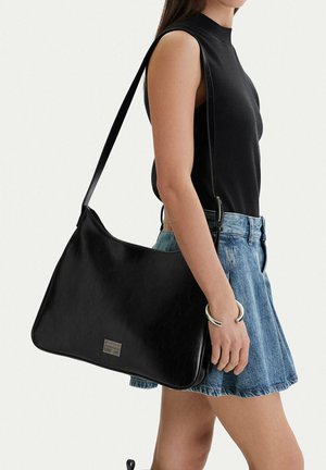 Mujer con blusa negra sin mangas y falda de mezclilla, llevando un bolso negro de hombro con una etiqueta plateada y usando una pulsera plateada.