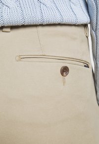 Béžové kalhoty s hladkým povrchem, se zapínáním na knoflík, jemným švem a boční kapsou. Nad nimi je vidět světle modrý pletený svetr.