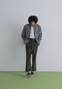 Geruite shirt in grijs en wit, witte tanktop, olijfgroene wijde broek, en bruine schoenen op een groene vloer. Minimalistische setting.
