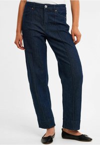 Dunkelblaue Jeanshosen mit geradem Schnitt, versehen mit Vordertaschen und rückseitigen Nähdetails. Getragen mit schwarzen Ballerinas.