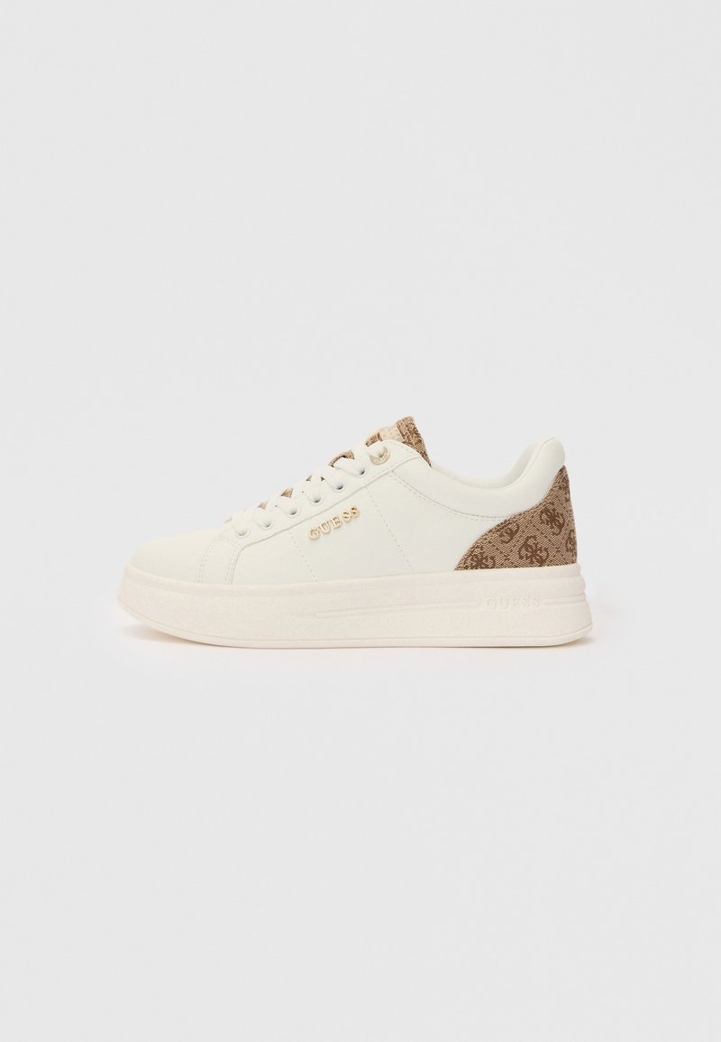 Matala valkoinen tennari, jossa on beige kuvioitu kantapää ja kieli, kultainen "GUESS"-logo kyljessä ja valkoiset nauhat valkoisella taustalla.