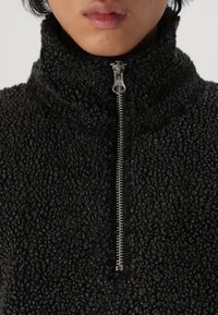 Sudadera de forro polar negra con cuello alto, que cuenta con una cremallera frontal, superficie texturizada y apariencia suave y acogedora.
