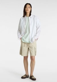Weißes Hemd mit Knöpfen und Kontrastnähten über einem hellgrünen T-Shirt, kombiniert mit beigen Cargo-Shorts und gemusterten Slippern.