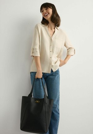 Kobieta w beżowej koszuli na guziki i niebieskich dżinsach trzyma dużą czarną torbę na ramię, uśmiechając się na jasnym, jednolitym tle.