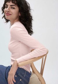 Helles rosa Langarmoberteil aus strukturiertem Strick, kombiniert mit dunkelblauen Jeans mit Taschen und metallischen Akzenten, auf einem Holzhocker sitzend.