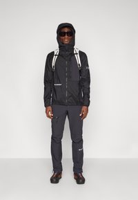 Zwarte waterdichte jas met een rits aan de voorkant en een capuchon, gecombineerd met donkere broeken en multi-kleurige, robuuste schoenen. Reflecterende rugzakbanden aanwezig.