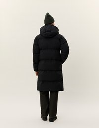 Persona che indossa un lungo piumino nero con cappuccio, pantaloni scuri, scarpe nere e un berretto verde oliva, in piedi con le spalle rivolte.