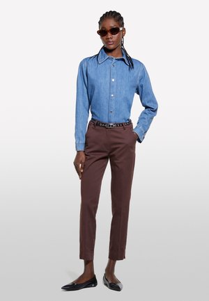 Camicia di jeans celeste con bottoni, pantaloni marroni su misura, ballerine nere a punta e occhiali da sole marroni con montatura sottile.