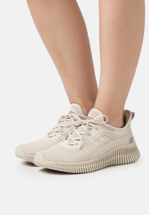 Pieds portant des baskets en tissu mesh beige avec semelles rainurées et lacets sur un fond clair.