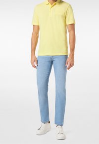 Man med gul kortärmad polotröja, ljusblå slim-fit jeans och vita snörskor som står framför en vit bakgrund.