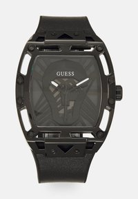 Must друзей дизаин черная Guess часовидет с прямоугольным корпусом с вырезанными деталями, текстурированным черным кожаным ремешком и узорчатым темным циферблатом с серебряными стрелками.