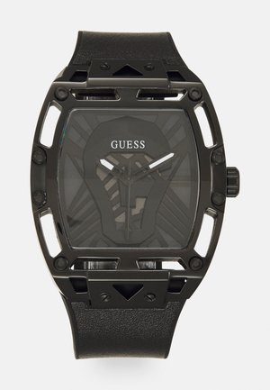 Montre Guess noire avec un boîtier rectangulaire et des découpes, bracelet en cuir noir texturé et un cadran sombre à motifs avec des aiguilles argentées.