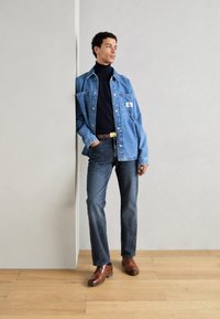 Denim jacket in light blue with a patch, black turtleneck, dark jeans, and brown leather shoes, standing against a neutral wall.