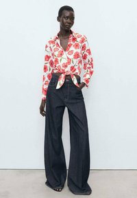 Camicia con stampa floreale rossa e verde su sfondo bianco, legata in vita. Abbinata a jeans in denim blu scuro a gamba larga.