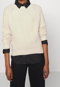 Krämfärgad stickad tröja med ribbade ärmslut och nederkant, lagd över en svart skjorta med krage. Tröjan har ett strukturerat mönster och raglanärmar.