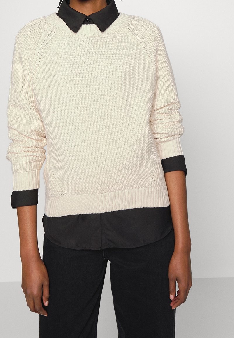 Krämfärgad stickad tröja med ribbade ärmslut och nederkant, lagd över en svart skjorta med krage. Tröjan har ett strukturerat mönster och raglanärmar.