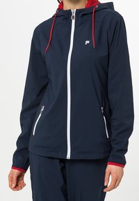 Marinblå vattentät jacka med zip-fram, röda detaljer på huva och ärmmuddar, två fickor med dragkedja och vita dragkedjedetaljer.