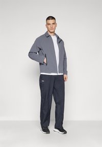 Gray zip-up jacket with front pockets, paired with dark navy pants featuring a logo. Model stands in a neutral background, wearing black shoes.