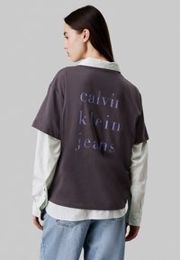 Camiseta gris de gran tamaño con "calvin klein jeans" impreso en púrpura claro, superpuesta sobre una camisa blanca con collar, y combinada con jeans azul claro.