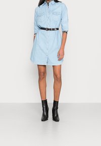 Svetle modrá džínová šaty košile s límcem, náprsními kapsami, knoflíkovým zapínáním a černým koženým páskem; v kombinaci s černými texturovanými kotníkovými botami.