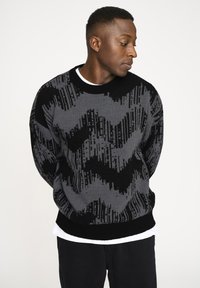 Strickpullover in Schwarz und Grau mit einem Zickzackmuster. Besitzt einen runden Ausschnitt und Bündchen, über einem weißen T-Shirt getragen, mit einem lockeren Schnitt.