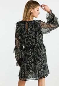 Schwarzes Kleid mit floralem Muster, durchsichtigen, rüschenbesetzten Ärmeln, taillierter Passform und einem tiefen Schlüssel-Ausschnitt auf dem Rücken. Leichter, strukturierter Stoff.