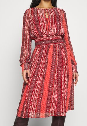 Femme portant une robe midi rouge à motifs avec manches longues, encolure goutte d'eau et taille cintrée, associée à des bottes foncées.