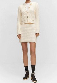 Cardigan en maille crème avec boutons à l'avant et deux poches, associé à une jupe ajustée assortie ; porté avec des chaussettes noires et des chaussures à talons dorés.