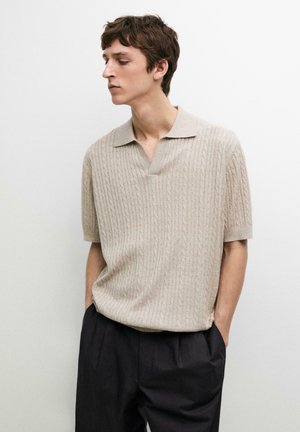 Giovane uomo che indossa una polo lavorata a maglia beige a maniche corte e pantaloni neri plissettati, in piedi con le mani nelle tasche contro una parete bianca.