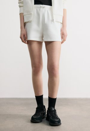 Kobieta w białych szortach z troczkiem, czarnych skarpetkach i czarnych trampkach, stojąca na szarym podłożu na tle białej ściany.