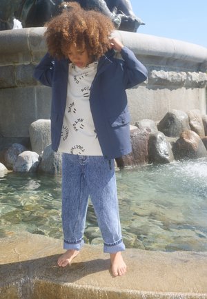 Ein Kind trägt eine marineblaue wasserabweisende Jacke über einem weißen Hemd mit schwarzen Textmustern und hellblauen Jeans, steht barfuß am Wasser.