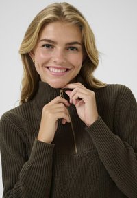 Hnědý svetr na zip s vysokým límcem, žebrovanou texturou a zlatým zipem, který nosí osoba s dlouhými vlasy.