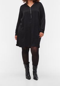 Vestito nero con colletto a zip, maniche lunghe e vestibilità ampia, realizzato in tessuto liscio, abbinato a stivali neri con punta affilata.