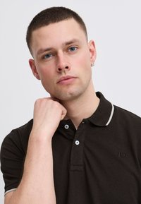 Tmavo hnedá polo košeľa s golierom, svetlosivé akcenty, dve gombíky a malý detail loga na ľavej strane hrude. Model pózuje s rukou pri brade.