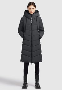 Mustal puffer-jakk pikendatud pikkus, kapuutsiga, ševronmuster, esi-zipper sulgemine, nööriga, ja nähtav logo detail zipperi peal.